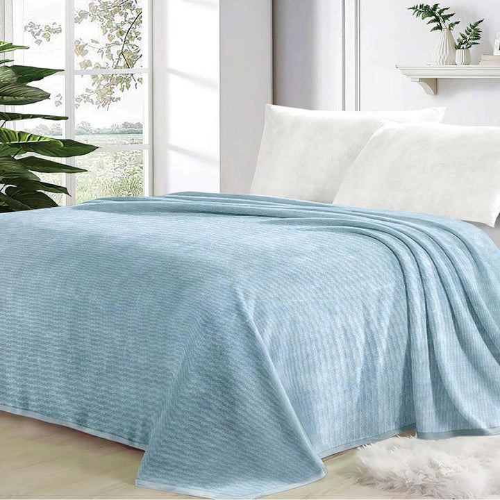 Light blue textured blanket draped over a bed in a bright bedroom with plants.
