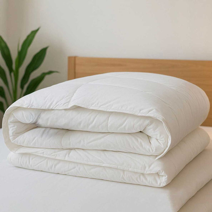 Folded white quilt on a bed with a wooden headboard and plant in the background