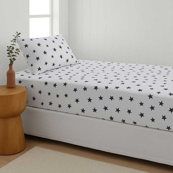 Bed with star-patterned sheet set in a room with a wooden side table and plant.