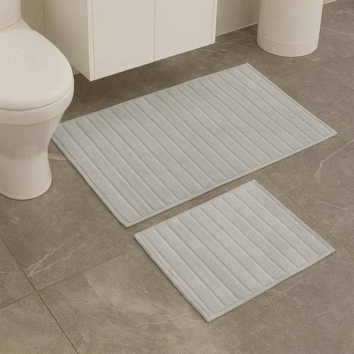 Two light gray bath mats on a bathroom floor with a toilet and sink in the background.