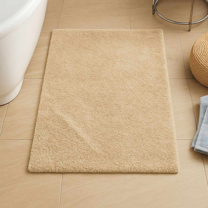 Beige bath mat on a tiled bathroom floor with a stool and towel in the background.