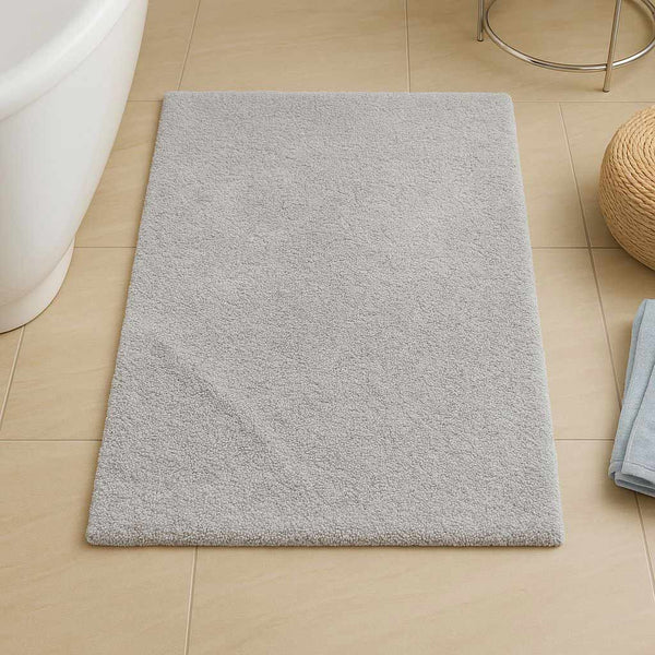 Gray bath mat on a bathroom floor with a towel and rug in the background