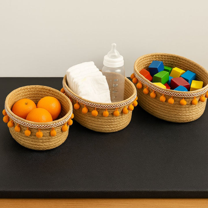 Three woven baskets with pom-pom details on a black surface, containing oranges, baby bottle, and colorful blocks.