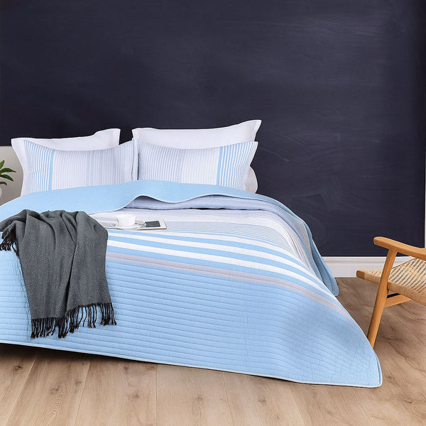 Light blue and white summer bedspread with vertical pinstripes, styled on a bed with matching pillows, a dark gray throw, and a navy accent wall