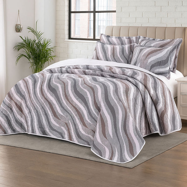 Wavy patterned summer bedspread in soft brown, grey, and mauve tones with matching shams, styled in a light-filled bedroom with brick walls