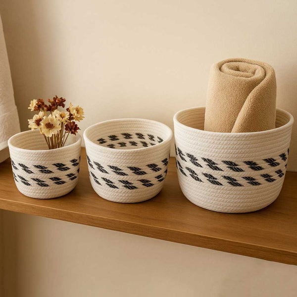 Three woven baskets with black pattern on a wooden shelf, one containing flowers and another a folded beige blanket.