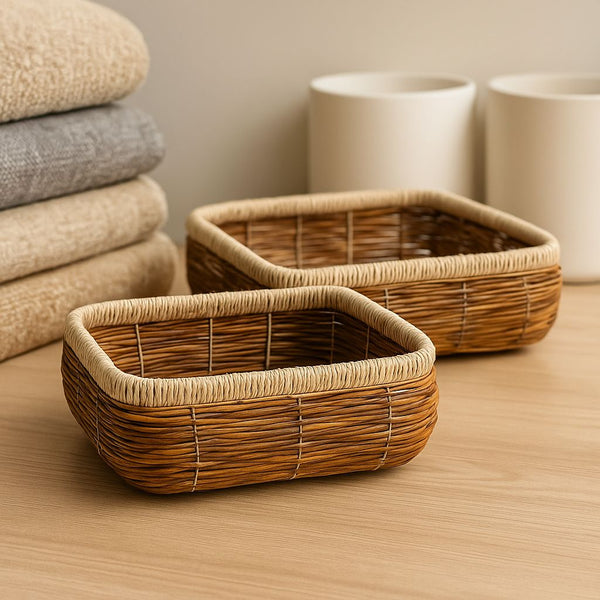 Two woven baskets on a wooden surface with stacked fabric and ceramic items in the background.